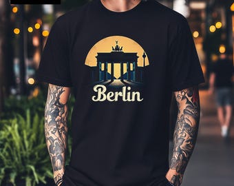 May include: A black short-sleeve t-shirt featuring a graphic print of the Brandenburg Gate in Berlin. The design includes a yellow circular sun background and the word Berlin in a stylized white font. The shirt has a crew neckline and a straight hem.