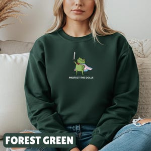May include: Forest green sweatshirt featuring a cartoon frog with a sword and cape, with the text "PROTECT THE DOLLS". The sweatshirt is a solid forest green color.