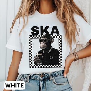 May include: White t-shirt with the word "SKA" in black block letters. The shirt features a graphic of a monkey wearing sunglasses and a black polo shirt, holding a glass of dark beer, set within a black and white checkered frame.