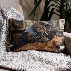 May include: A rectangular cushion with a humorous illustration of two cats in a tattoo parlour. One cat, wearing a denim jacket and cap, tattoos the other cat. The pillow has a neutral background.