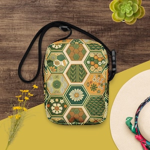 May include: A crossbody bag with a black strap and a geometric pattern. The bag features a repeating pattern of green, orange, and beige hexagons with floral and leaf designs. The bag is rectangular with rounded corners.