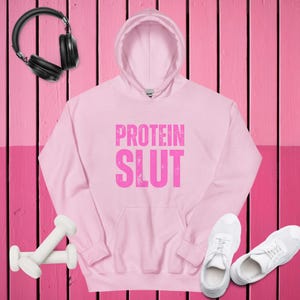 Könnte beinhalten: Hellrosa Kapuzenpullover mit dem Text "PROTEIN SLUT" in rosa. Das Bild zeigt auch schwarze Kopfhörer, weiße Kurzhanteln und weiße Turnschuhe, alles vor einem rosa Hintergrund.