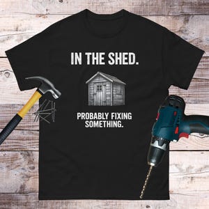 May include: Black t-shirt with the text "IN THE SHED. PROBABLY FIXING SOMETHING." above and below a graphic of a wooden shed. A hammer and drill are on either side of the shirt.