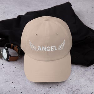 May include: A beige baseball cap featuring the word "ANGEL" embroidered in white, with white wing designs. The cap is made of a soft material and has a curved peak. A watch with a brown strap is visible in the background.