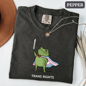 Puede incluir: Camiseta color pimienta con una rana de dibujos animados con una espada, pajarita y capa de orgullo trans. El texto "TRANS RIGHTS" está impreso debajo. También se ve un collar plateado con un colgante.