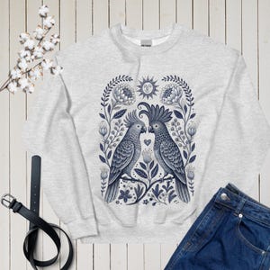 May include: A pale grey sweatshirt with a blue and white illustration of two cockatoos, floral elements, and a sun design. The sweatshirt is styled with blue jeans and a black belt.