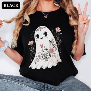 Può includere: Una t-shirt nera con la grafica di un fantasma bianco decorato con fiori rosa, foglie e un piccolo cuore rosso. Il fantasma ha grandi occhi neri e una bocca sorridente. La parola BLACK è stampata in bianco nell'angolo in alto a sinistra.