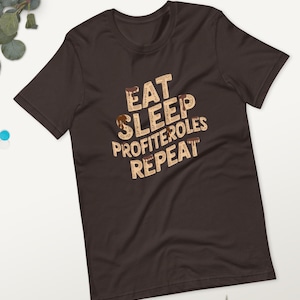Pode incluir: Uma t-shirt castanha escura com as palavras "EAT SLEEP PROFITEROLES REPEAT" em castanho claro, com textura e detalhes de chocolate a pingar. A t-shirt tem gola redonda e mangas curtas.
