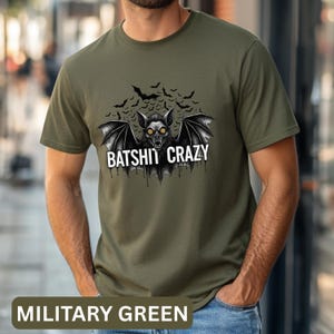 Puede incluir: Camiseta verde militar con un gráfico de murciélago y el texto "BATSHIT CRAZY". El murciélago tiene ojos amarillos y está rodeado de siluetas de murciélagos más pequeñas. El color de la camiseta está etiquetado como "MILITARY GREEN" en la parte inferior.