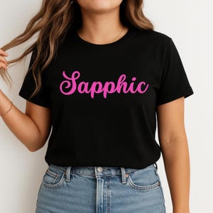 Puede incluir: Camiseta negra con la palabra "Sapphic" en rosa brillante, con fuente cursiva. La camiseta se combina con unos vaqueros azules. La modelo lleva una pulsera dorada.
