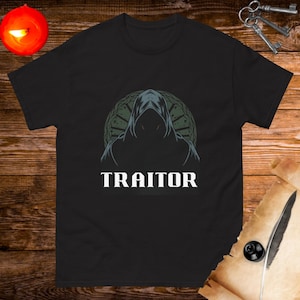 May include: Black t-shirt featuring a graphic of a hooded figure in front of a circular design. The text "TRAITOR" is printed below the figure.