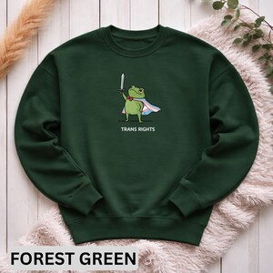 May include: Forest green sweatshirt featuring a cartoon frog holding a sword and wearing a cape with the transgender flag colors. The text "TRANS RIGHTS" is below the frog. The sweatshirt is displayed on a neutral-colored surface.