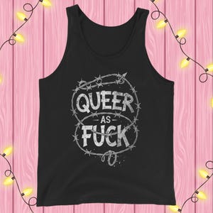 Puede incluir: Camiseta sin mangas negra con el texto "QUEER AS FUCK" en letras blancas desgastadas, rodeado por un diseño de alambre de púas. La camiseta sin mangas está hecha de un material suave y es adecuada para uso casual.
