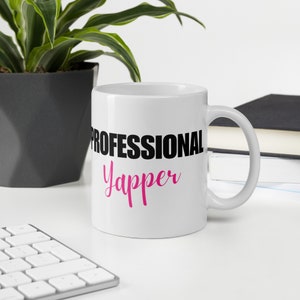 Pode incluir: Caneca de café de cerâmica branca com texto preto que diz "Professional Yapper" em uma fonte cursiva rosa.
