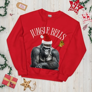 Könnte beinhalten: Ein rotes Langarm-Sweatshirt mit einer Grafik eines Gorillas, der eine rot-weiße Weihnachtsmütze trägt. Über dem Gorilla ist der Text JUNGLE BELLS in Weiß gedruckt. Rechts neben dem Gorilla befindet sich eine kleine goldene Glockengrafik.
