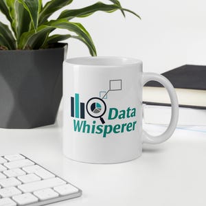 May include: White ceramic mug with the words "Data Whisperer" in teal and black. The mug features a graphic of a magnifying glass, pie chart, and bar graph. The mug is on a white surface.