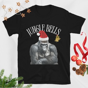 Könnte beinhalten: Ein schwarzes T-Shirt mit kurzen Ärmeln und einem Aufdruck eines Gorillas, der eine rot-weiße Weihnachtsmütze trägt. Über dem Gorilla steht der Text JUNGLE BELLS in Weiß. Rechts neben dem Kopf des Gorillas befindet sich eine kleine goldene Glocke.