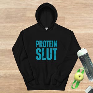 Könnte beinhalten: Schwarzer Kapuzenpullover mit dem Text "PROTEIN SLUT" in Blau.