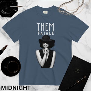 May include: Midnight blue t-shirt featuring the text "THEM FATALE" in white. The graphic is a monochrome image of a person in a hat and dress, with a finger to their lips. A black notebook and lipstick are also visible.