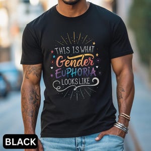 以下が含まれることがあります： 「THIS IS WHAT Gender EUPHORIA LOOKS LIKE」というカラフルな文字がプリントされた黒いTシャツ。デザインには星と装飾的な線が含まれています。画像の下部には「BLACK」という文字が印刷されています。