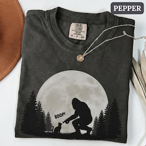 May include: A charcoal grey crew neck t-shirt featuring a graphic of a large moon, a forest silhouette, and a Bigfoot figure reaching out to touch a small cat with the text BOOP! printed above the cat. The shirt has a Comfort Colors brand label.