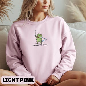 May include: Light pink sweatshirt featuring a cartoon frog holding a sword and wearing a cape with the text "PROTECT THE DOLLS". The frog has a heart on its chest. The words "LIGHT PINK" are at the bottom of the image.
