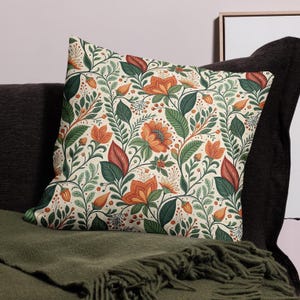 Może przedstawiać: Kwadratowa poduszka z kwiatowym wzorem. Wzór przedstawia pomarańczowe i czerwone kwiaty, zielone liście i małe pomarańczowe pąki na kremowym tle. Poduszka leży na ciemnozielonym kocu.