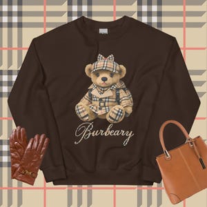 Puede incluir: Una sudadera marrón con un oso de peluche con sombrero y abrigo a cuadros. La palabra "Burbeary" está escrita en cursiva debajo del oso. En la imagen también hay guantes de cuero marrones y un bolso marrón.