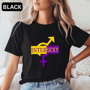 May include: A black short-sleeved t-shirt featuring a graphic design with the text INTERSEXY. The design includes a yellow and purple rectangular block overlaying a yellow and purple intersex symbol, with the word INTERSEXY printed in yellow and purple.