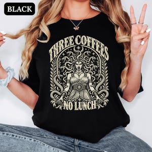 May include: A black t-shirt featuring a cream-coloured graphic print of Medusa with snakes for hair, surrounded by an archway of leaves and stars. The text "THREE COFFEES" is arched above the figure, and "NO LUNCH" is printed in a straight line below.