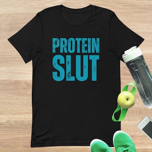 Pode incluir: Camiseta preta com o texto "PROTEIN SLUT" em letras turquesa desgastadas. A imagem inclui uma garrafa de água, uma maçã verde com uma faixa verde, um smartphone e tênis verdes.