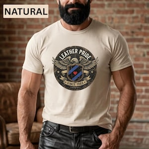 Op de afbeelding: Een naturelkleurig t-shirt met een cirkelvormige afbeelding van een adelaar, een schild met een blauwe streep en een rood hart, en de tekst "LEATHER PRIDE SINCE 1989". Het ontwerp is gecentreerd op de borst van het t-shirt met korte mouwen.