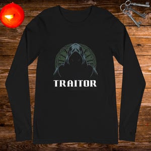 May include: Black long-sleeved shirt featuring a hooded figure against a circular design with the word "TRAITOR" in white lettering.