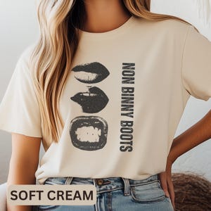 Puede incluir: Camiseta de color crema suave con un gráfico negro de tres juegos de labios y el texto "NON BINNY BOOTS" en negro. Las palabras "SOFT CREAM" están impresas en la parte inferior de la camiseta.