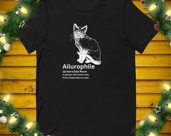 May include: Black t-shirt featuring a white illustration of a cat. The word "Ailurophile" is printed below the cat, along with a definition: "A person who loves cats."