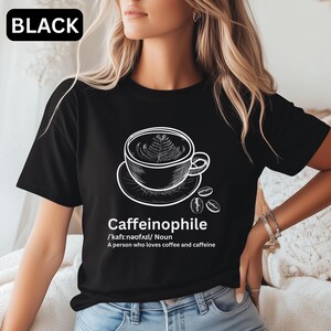 May include: Black t-shirt featuring a white illustration of a coffee cup with latte art, coffee beans, and the word "Caffeinophile" with its pronunciation and definition. The shirt has a crew neck.