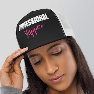 Peut inclure: Un chapeau de camionneur noir et blanc avec un dos en maille blanche. Le devant du chapeau porte le texte "PROFESSIONAL Yapper" en lettres blanches avec le mot "Yapper" en rose.