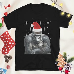 Könnte beinhalten: Ein schwarzes Kurzarm-T-Shirt mit einer Grafik eines grauen Gorillas, der eine rot-weiße Weihnachtsmütze trägt. Der Gorilla ist mittig auf der Brust platziert und von weißen vierzackigen Sternen auf dem schwarzen Stoff umgeben.