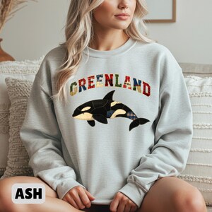 Pode incluir: Camisola cinza-cinza com a palavra "GREENLAND" num design de patchwork no peito. Abaixo do texto, uma orca preta e branca com detalhes de patchwork. A camisola é de gola redonda.