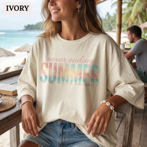 Könnte beinhalten: Elfenbeinfarbenes Langarmshirt mit dem Text "never ending SUMMER" in Pastellfarben. Das Shirt hat einen Rundhalsausschnitt und eine lockere Passform. Das Bild zeigt eine Person, die das Shirt trägt, mit einer Strandszene im Hintergrund.