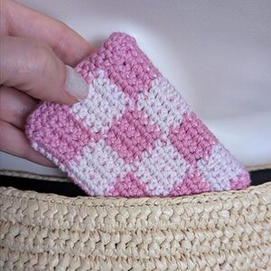 May include: A pink and white crocheted square is held above a woven basket. The square has a checkerboard pattern of pink and white. The basket is made of a light tan woven material.