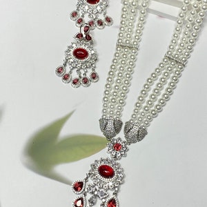 May include: A luxurious jewelry set featuring a multi-strand pearl necklace with a large pendant and matching earrings. The pendant and earrings have a silver-tone setting with a large red gemstone surrounded by smaller red and clear stones.