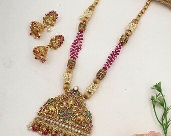 May include: A gold necklace with a large pendant featuring an intricate design of elephants, flowers, and gemstones. The necklace is made of pink and white beads and comes with a pair of matching earrings.