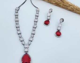 Puede incluir: Un collar y pendientes de plata con una piedra roja en forma de lágrima y piedras de circonita cúbica transparentes.