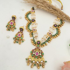 May include: An ornate gold-toned necklace and matching earrings set. The necklace features multiple strands of white beads, green and gold accents, and a large pendant with pink and green gemstones. The earrings mirror the pendant design.