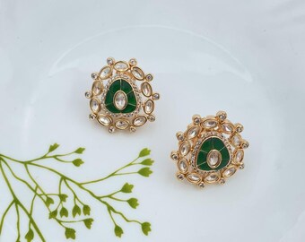 May include: A pair of gold-toned stud earrings. Each earring features a central green stone surrounded by clear, oval-shaped crystals. The earrings are set on a white surface with a green floral accent.