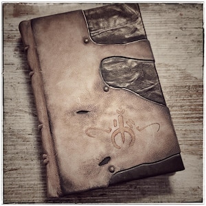May include: A brown leather-bound journal with a decorative design on the front cover. The journal has a dark brown leather flap that wraps around the front cover and is secured with a metal clasp. The journal has a worn and aged look, suggesting it is an antique.