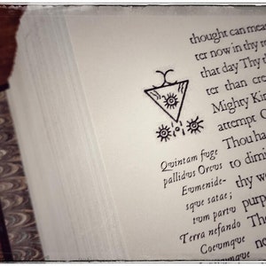 K&ouml;nnte beinhalten: Eine Seite aus einem Buch mit einer Schwarzwei&szlig;illustration eines Symbols mit einem Dreieck und Kreisen. Der Text auf der Seite lautet: "Quintam fuge pallidus Orcus Eumenide- sque satae; tum parto Terra nefando Cocumque mave".