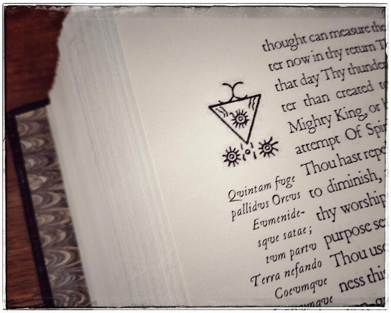 K&ouml;nnte beinhalten: Eine Seite aus einem Buch mit einer Schwarzwei&szlig;illustration eines Symbols mit einem Dreieck und Kreisen. Der Text auf der Seite lautet: "Quintam fuge pallidus Orcus Eumenide- sque satae; tum parto Terra nefando Cocumque mave".