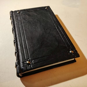 K&ouml;nnte beinhalten: Ein schwarzes, ledergebundenes Notizbuch mit dekorativen Metallnieten an den Ecken. Das Notizbuch hat einen strukturierten Einband und einen R&uuml;cken mit gepr&auml;gten Details. Die Seiten sind am Rand sichtbar.